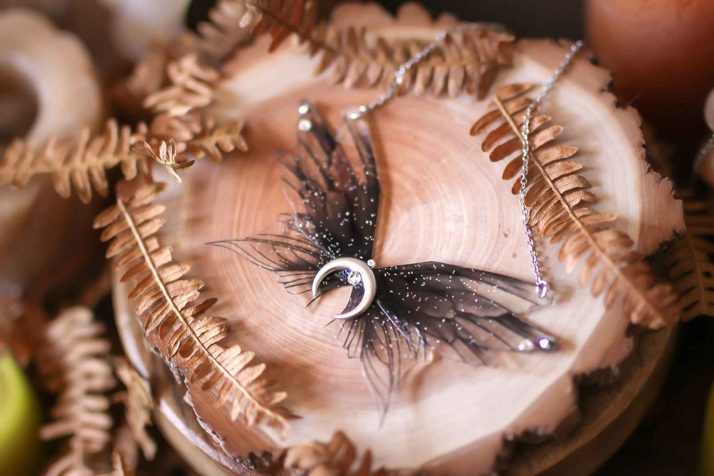 Collier Coven des Sorcières de la marque Faërie, affichant un design enchanteur avec quatre ailes de fées en dégradé noir et des paillettes irisées, parfait pour révéler votre magie intérieure.