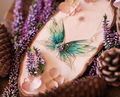 Lac scintillant - Collier orné de quatre Ailes de Fées aux teintes bleu profond et turquoise, décoré de paillettes irisées et d'une pierre bleue centrale, symbolisant la délicatesse et la beauté artisanale.