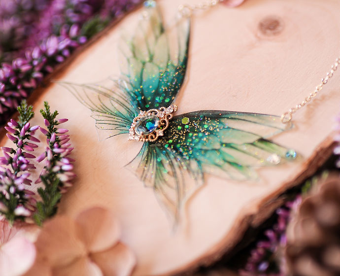 Découvrez le collier 'Lac scintillant' de Faërie, orné de quatre ailes de fées en dégradé bleu profond et turquoise, avec des paillettes irisées et un ornement en pierre bleue.