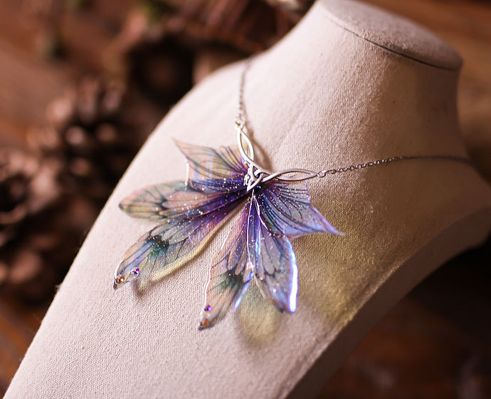 Collier féerique La Larme de l’Aube avec des Ailes de fée violettes et bleues scintillantes sous les reflets lumineux.