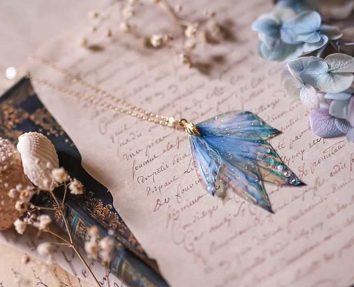 Collier Azurelle féerique avec quatre ailes de fée, inspirées des nageoires de sirènes, présentant un dégradé de bleu profond et clair, sublimé par des paillettes irisées.