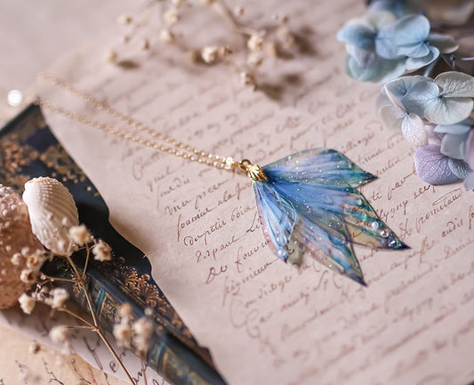 Collier Azurelle féerique avec quatre ailes de fée, inspirées des nageoires de sirènes, présentant un dégradé de bleu profond et clair, sublimé par des paillettes irisées.